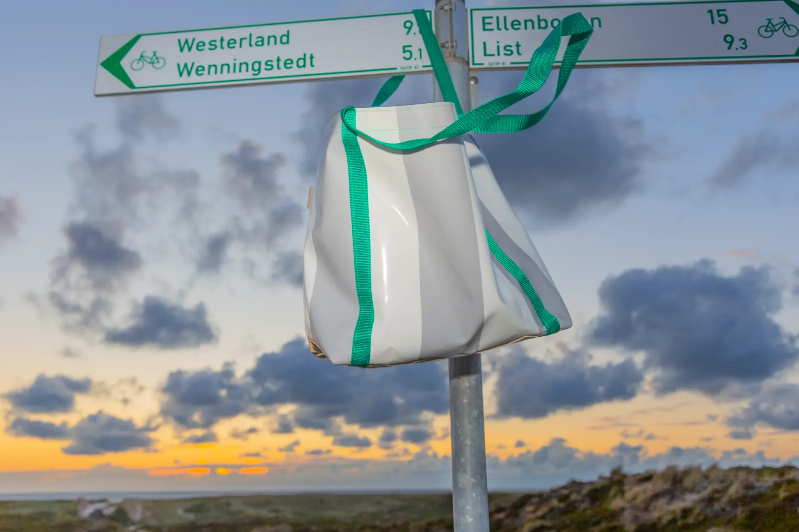 STURMflut Solo Shopper Bag aus original Sylter Strandkorbplane in Grau-Weiß mit individualisierbaren Trägerfarben am Sylter Fahrrad-Wegweiser Westerland–Wenningstedt vor Nordsee-Sonnenuntergang — handgefertigtes Unikat von STURMFREI, Fertigung auf Anfrage